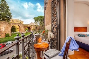 A balcony with a view of a historic bridge, a table with a drink, and a bed with a wooden headboard. - Hotel Emona Aquaeductus (Rome)