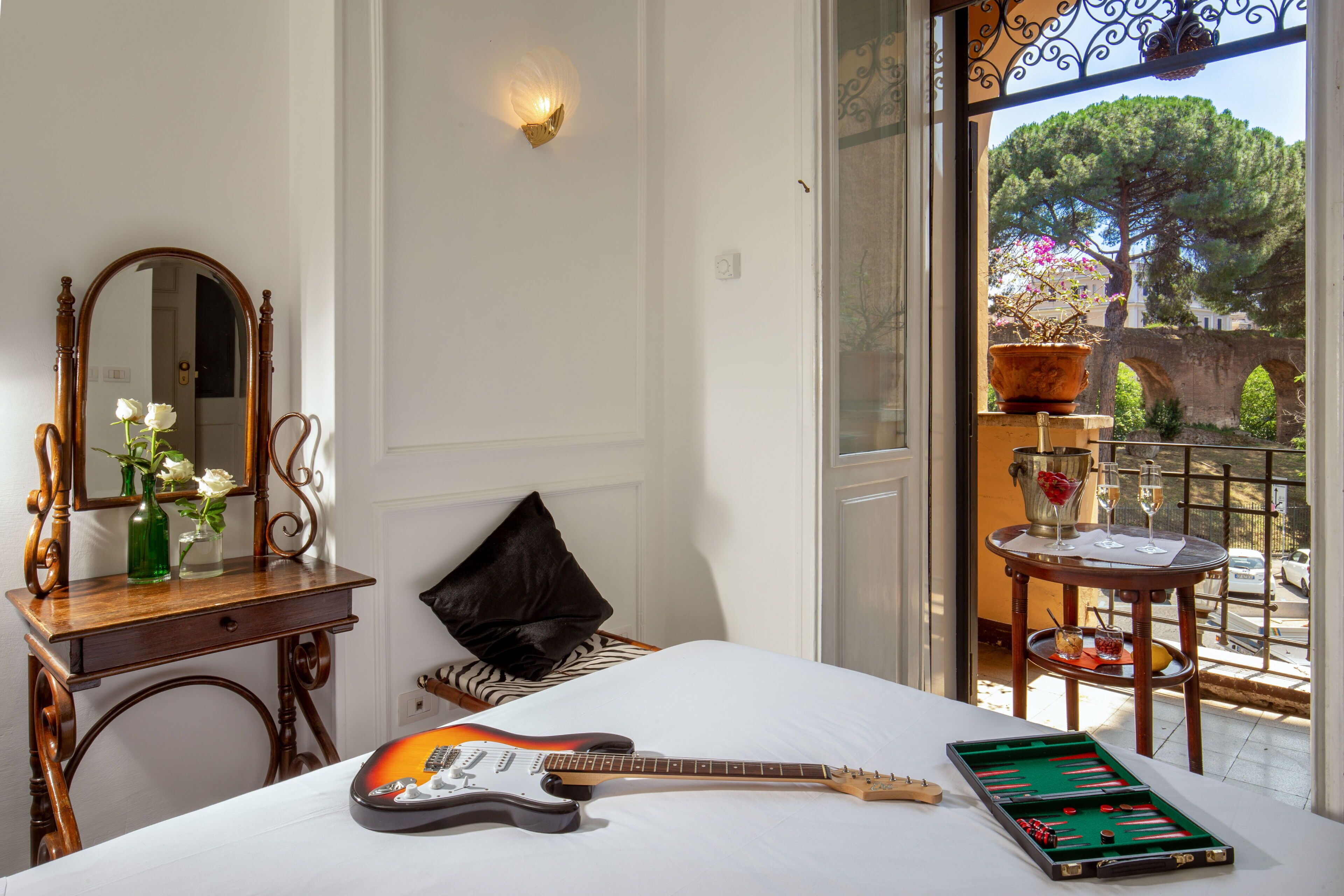 A room with a bed, a guitar, a green board game, a mirror, a vase with flowers, and a view of a tree and a bridge outside.