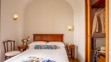 A neatly made bed with a wooden headboard, a bedside table with a lamp, a chair, and a wardrobe with a hanging garment.
