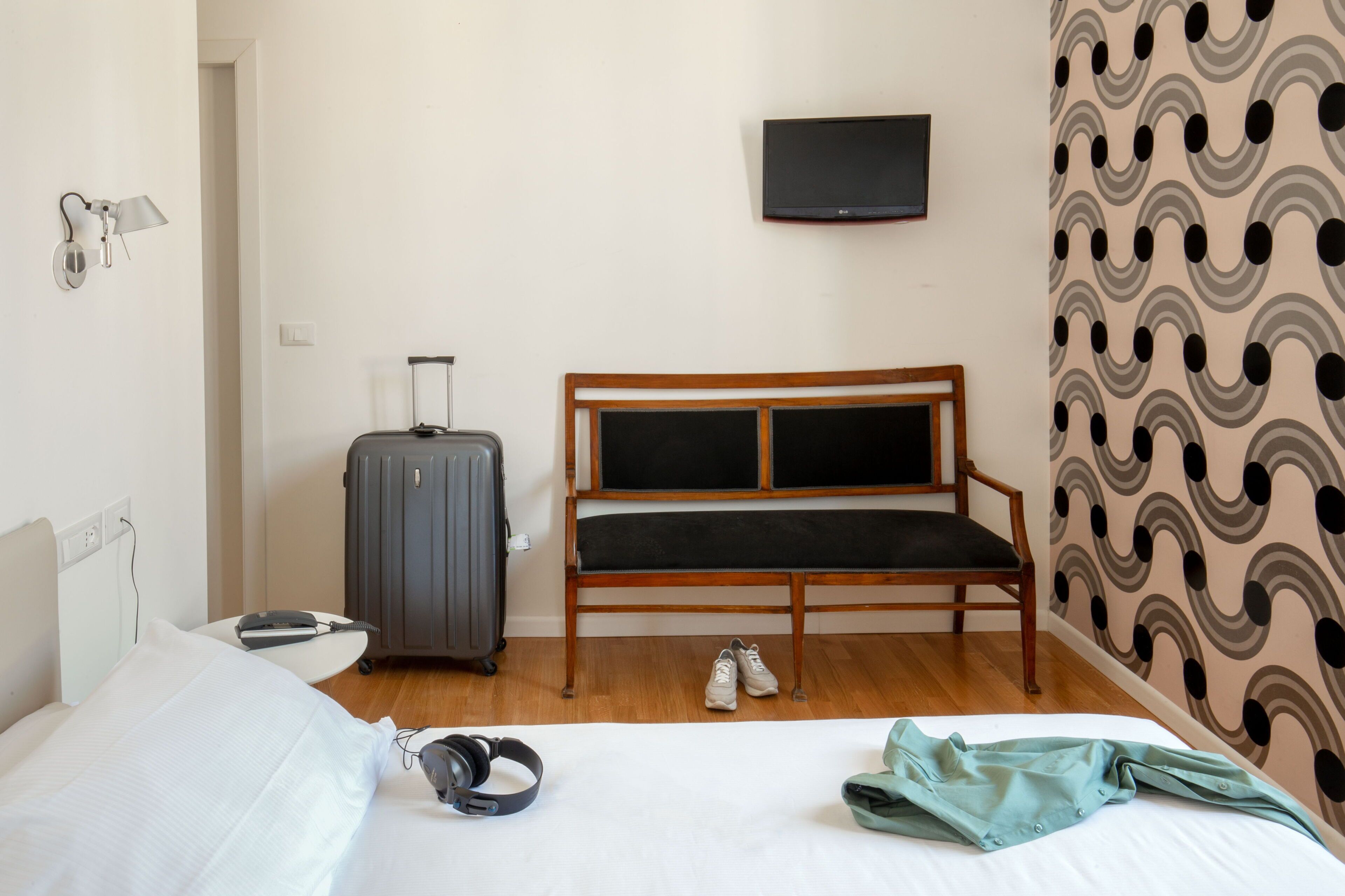 A hotel room with a bed, a wooden bench, a suitcase, a television, and patterned wallpaper.