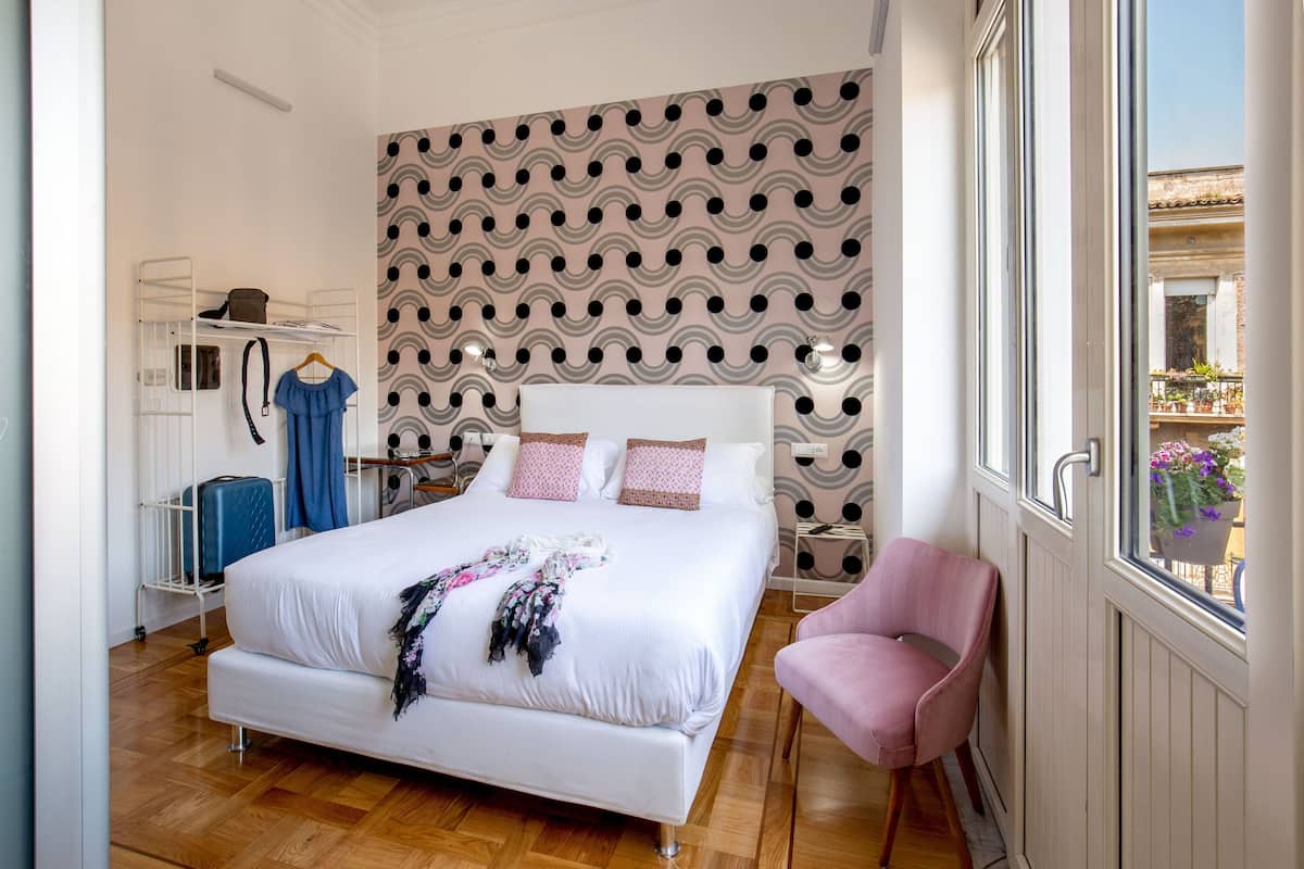 A bedroom with a large bed, a pink chair, a wardrobe, and a patterned wall.