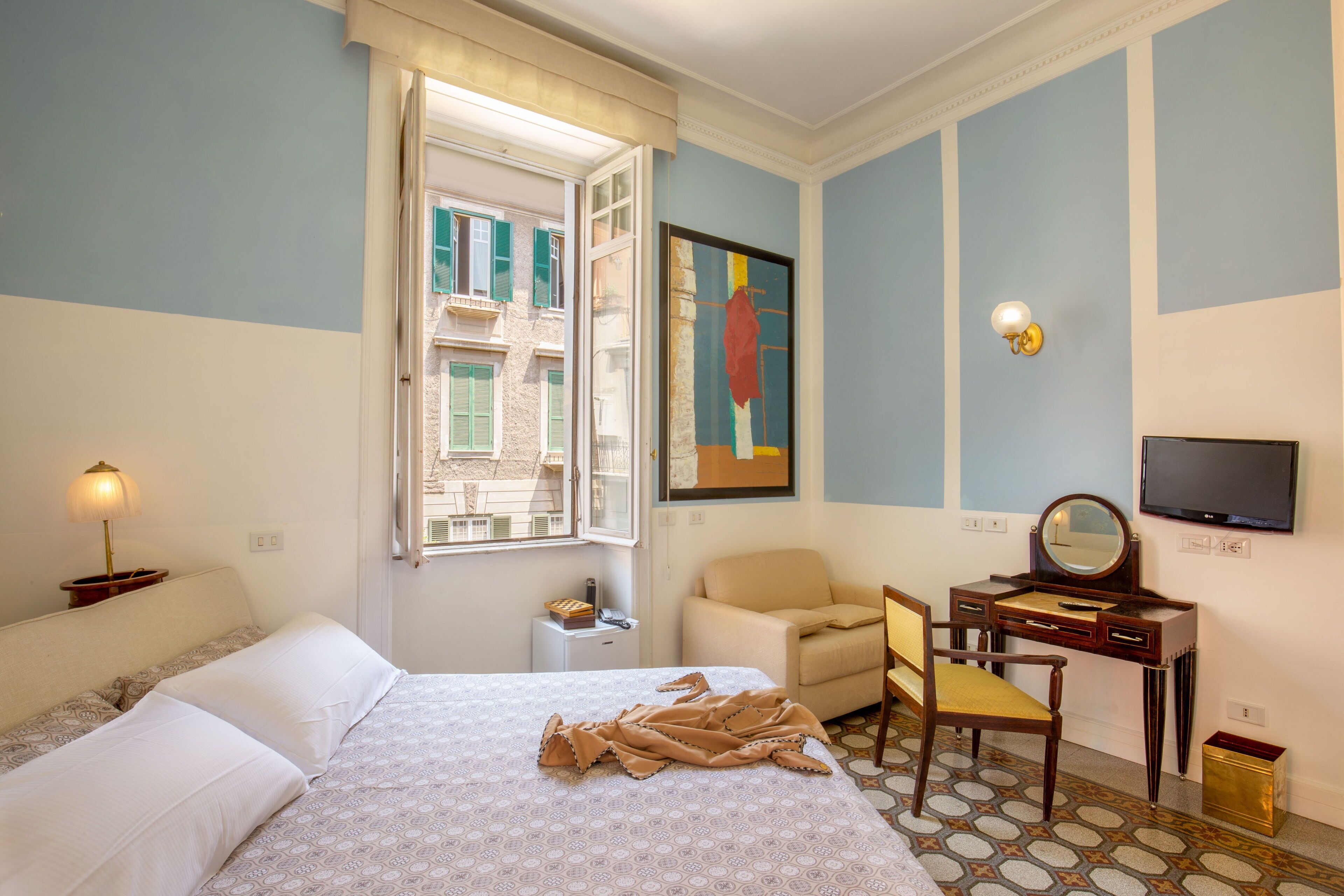 A hotel room with a bed, a desk with a mirror, a chair, a small table with a lamp, and a television on the wall.