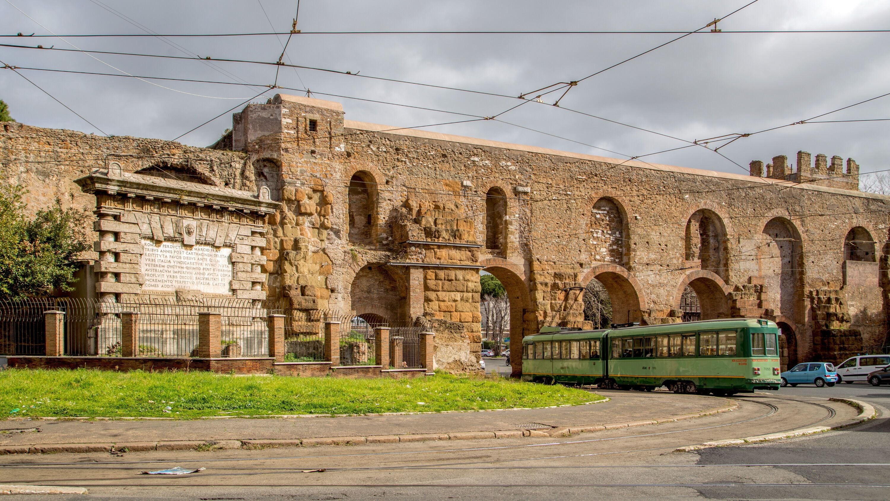Un antico edificio in pietra con numerose arcate e un tram verde in primo piano.