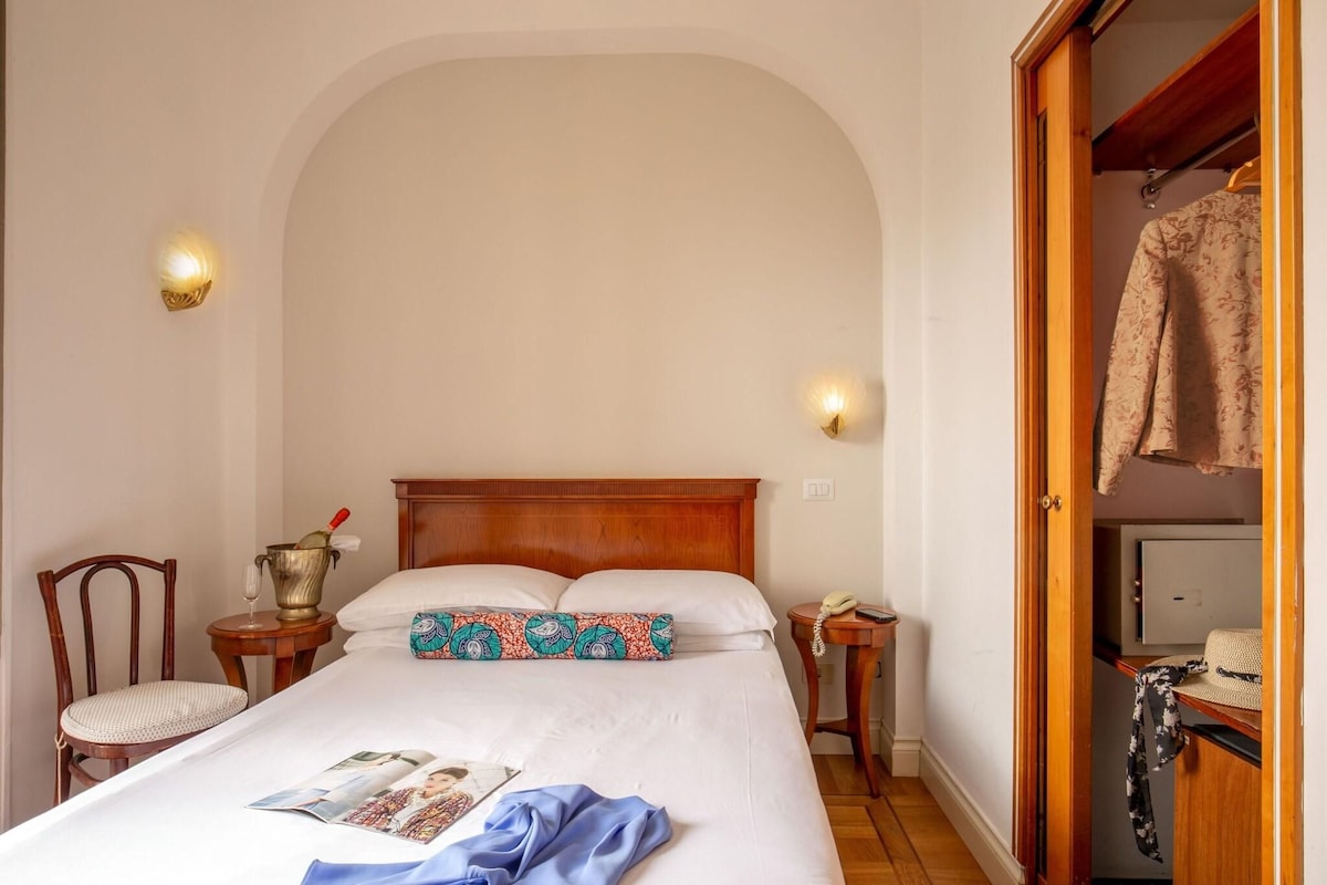 Un lit bien fait, avec une tête de lit en bois, une table de chevet avec une lampe, une chaise et une armoire où est suspendu un vêtement.