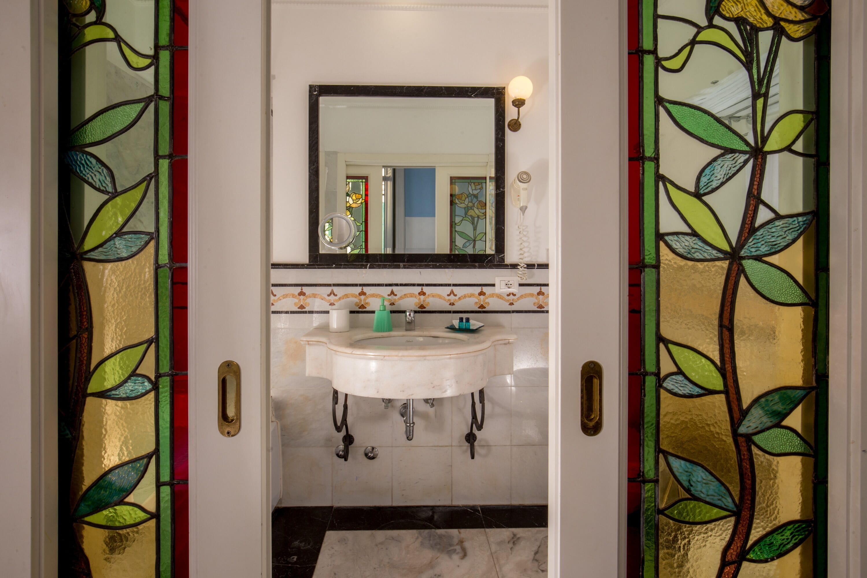 Een badkamer met een wit wastafelmeubel, een spiegel erboven en een decoratieve glas-in-looddeur.