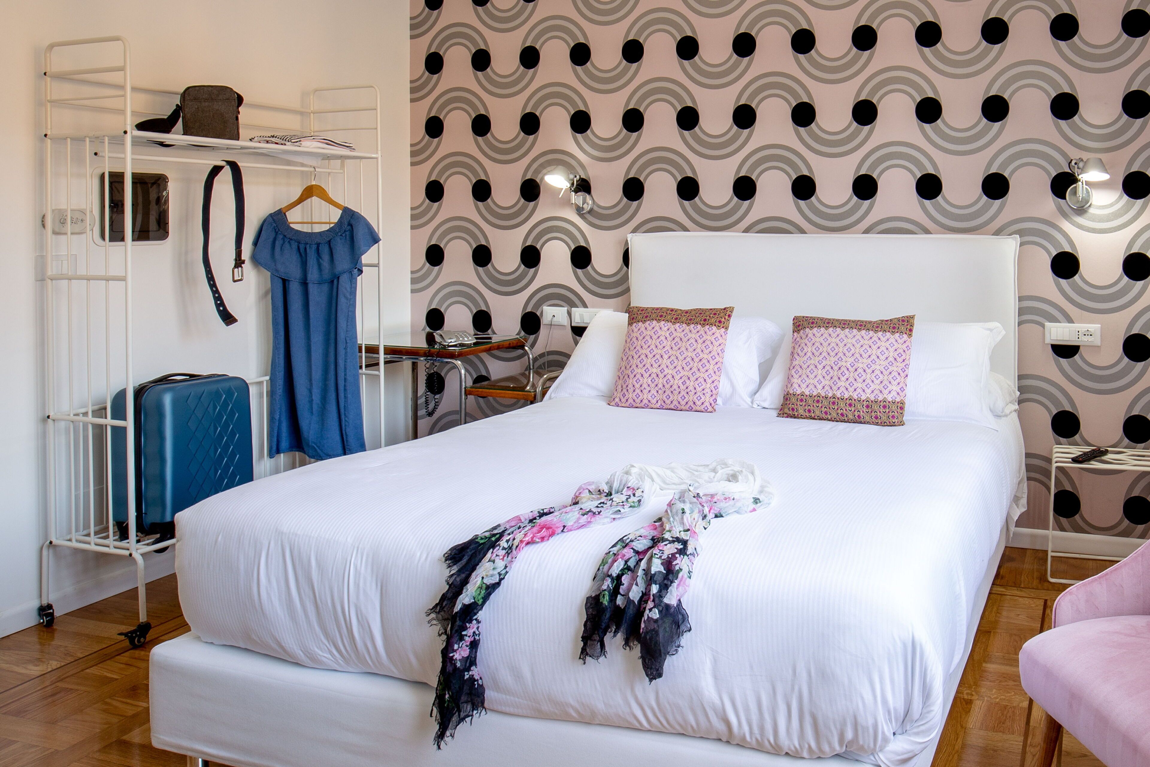 A neatly made bed with a floral throw, a suitcase, and a hanging dress on a metal shelf.