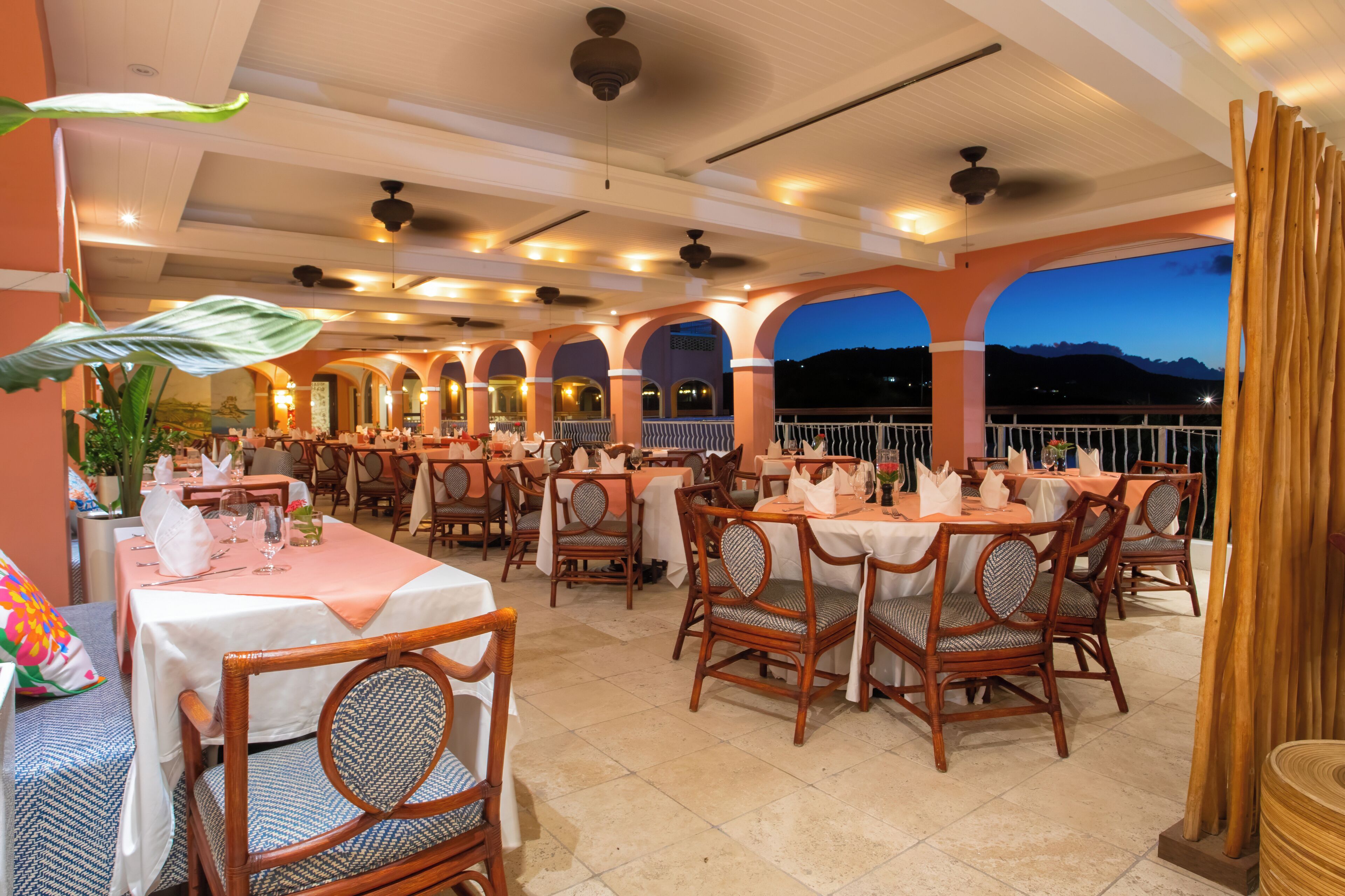 Un restaurante con varias mesas y sillas, un balcón con vistas a las montañas y un ventilador de techo.