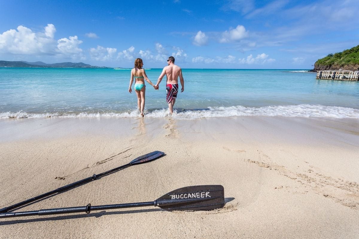 A beach with two people walking, a paddleboard, and a wooden structure on the right.