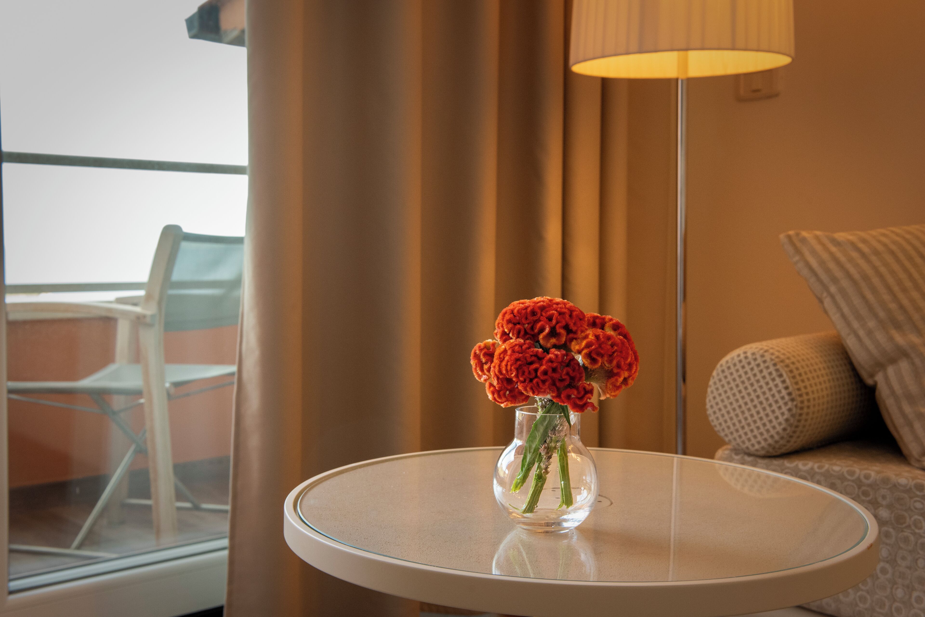 Ein runder Glastisch mit einer klaren Vase, in der Blumen, eine Lampe und ein Blick auf einen Balkon mit einem Stuhl sind.