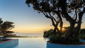 An infinity pool with a view of the sunset over the ocean, surrounded by trees and a bench.