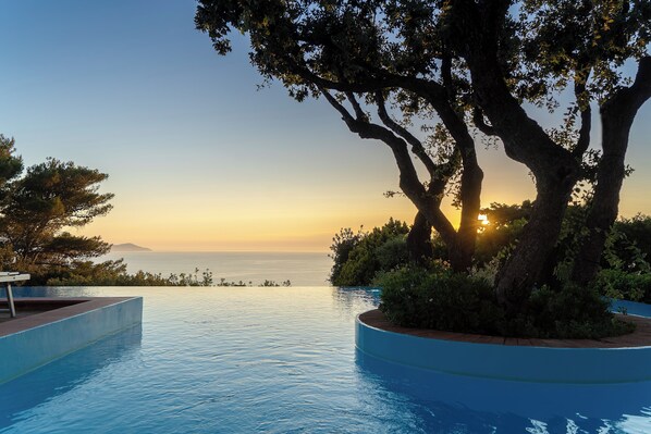 Una piscina infinita con vistas a la puesta de sol sobre el océano, rodeada de árboles y un banco.