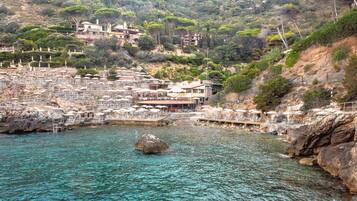 Un hotel sulla costa con posti a sedere all'aperto sul lungomare, un litorale roccioso e edifici su una collina.