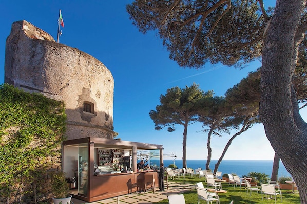 A coastal bar with outdoor seating, a stone tower, and a view of the sea.