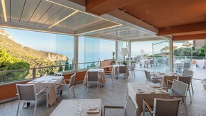 A restaurant with tables set for dining, large windows offering a view of the sea and hills, and a covered outdoor area.