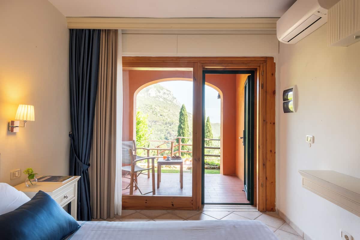 A hotel room with a bed, a nightstand, a lamp, a window with curtains, and an open door leading to a balcony with a table and chairs.
