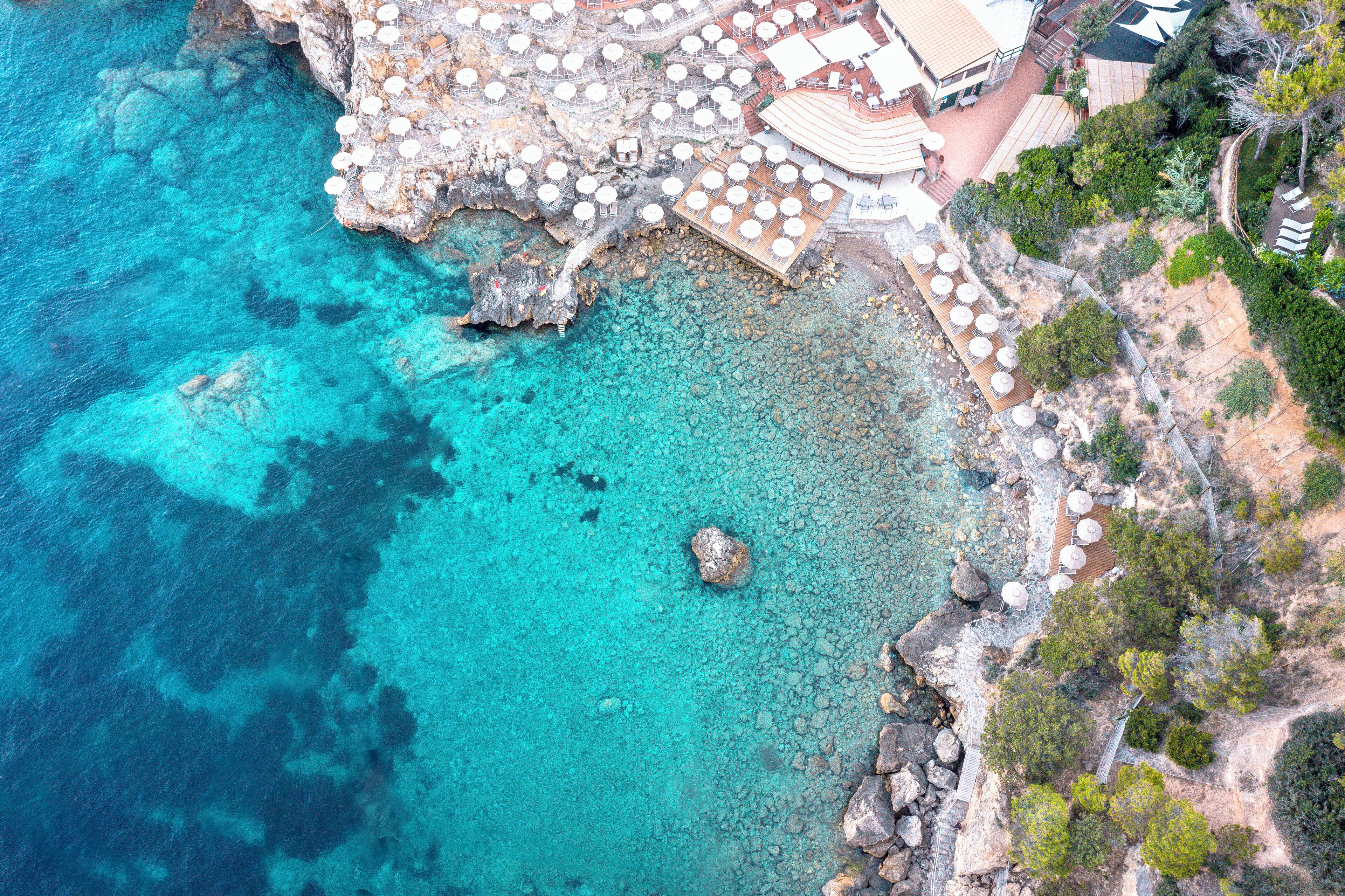 An aerial view of a coastal resort with clear turquoise waters, rocky terrain, and outdoor seating areas.