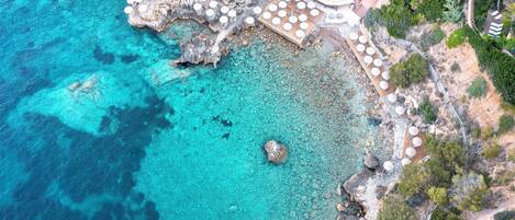 An aerial view of a coastal resort with clear turquoise waters, rocky terrain, and outdoor seating areas.