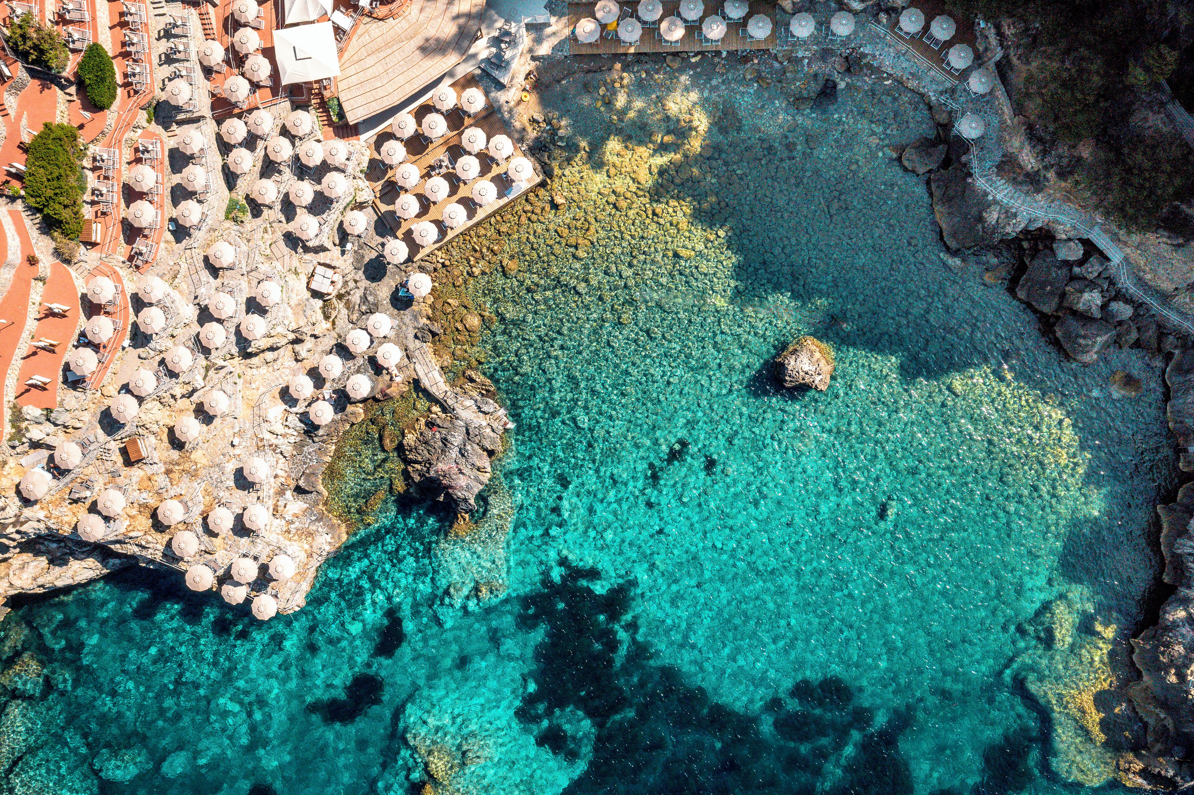 An aerial view of a coastal area with clear turquoise waters, rocky terrain, and a series of white umbrellas arranged in rows, likely indicating a beachside hotel or resort area.