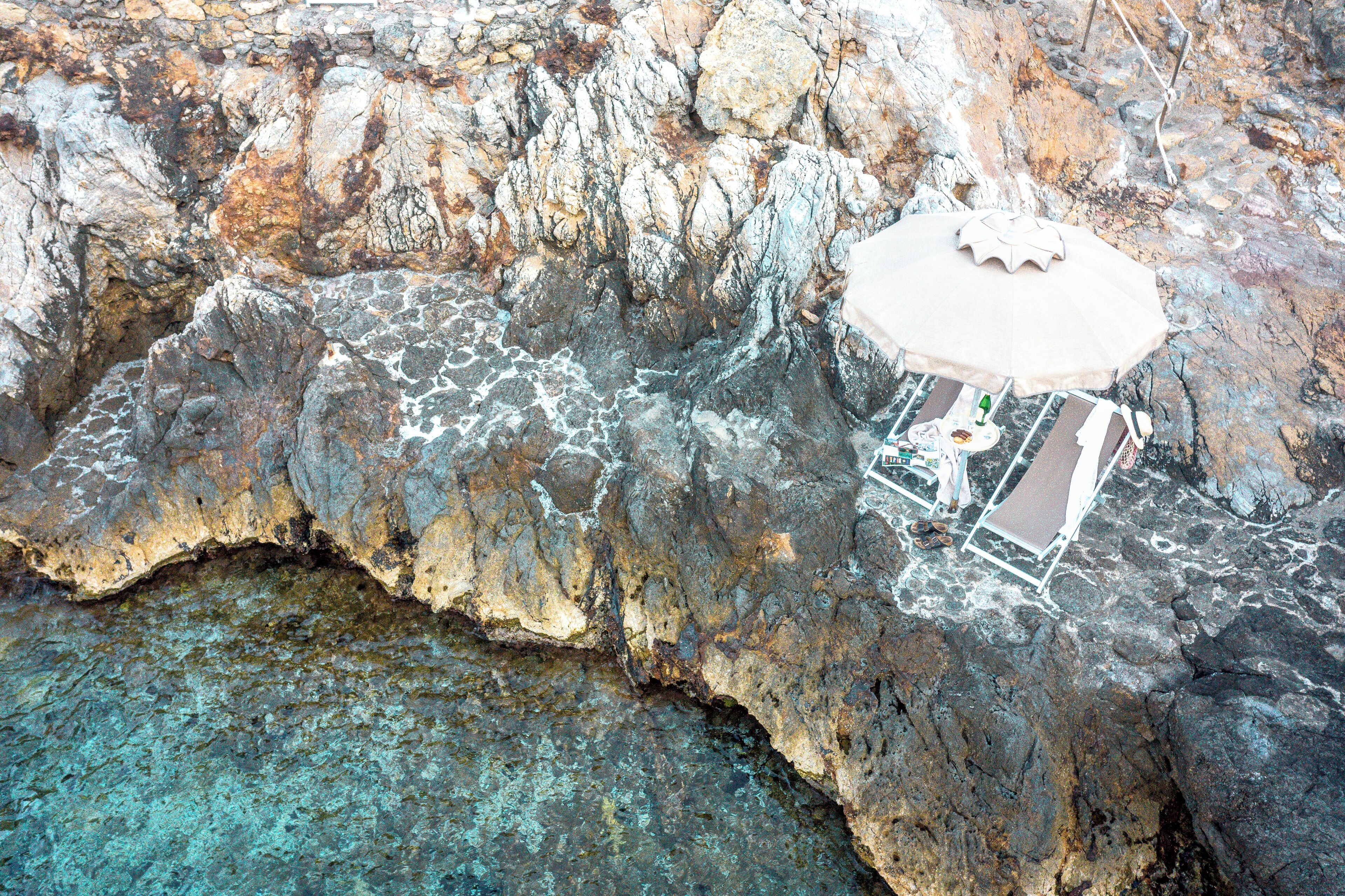 A rocky cliff with a small beach area featuring a single lounge chair and an umbrella.