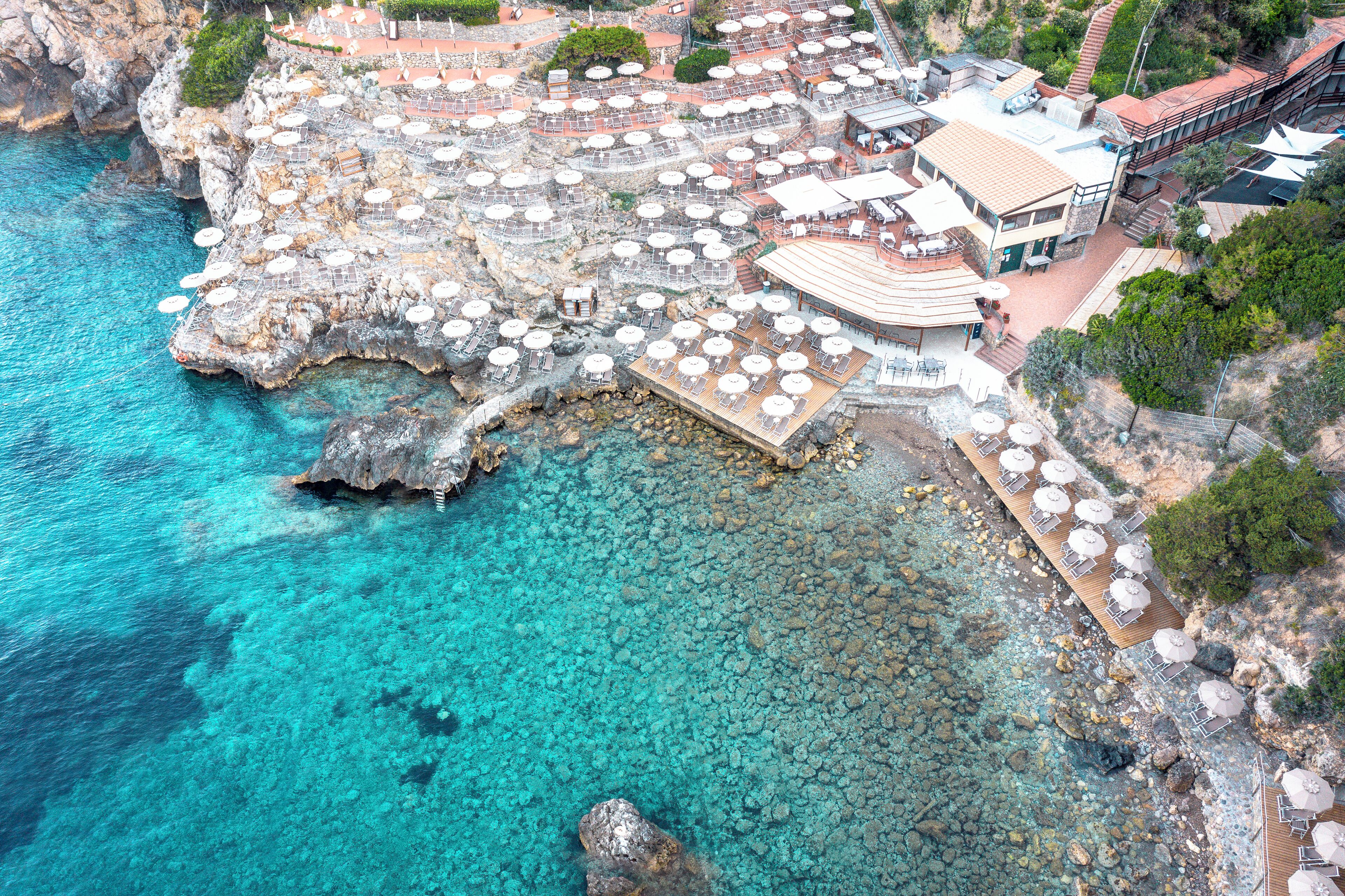 An aerial view of a coastal hotel with multiple sun loungers and umbrellas, a rocky shoreline, and clear turquoise water.