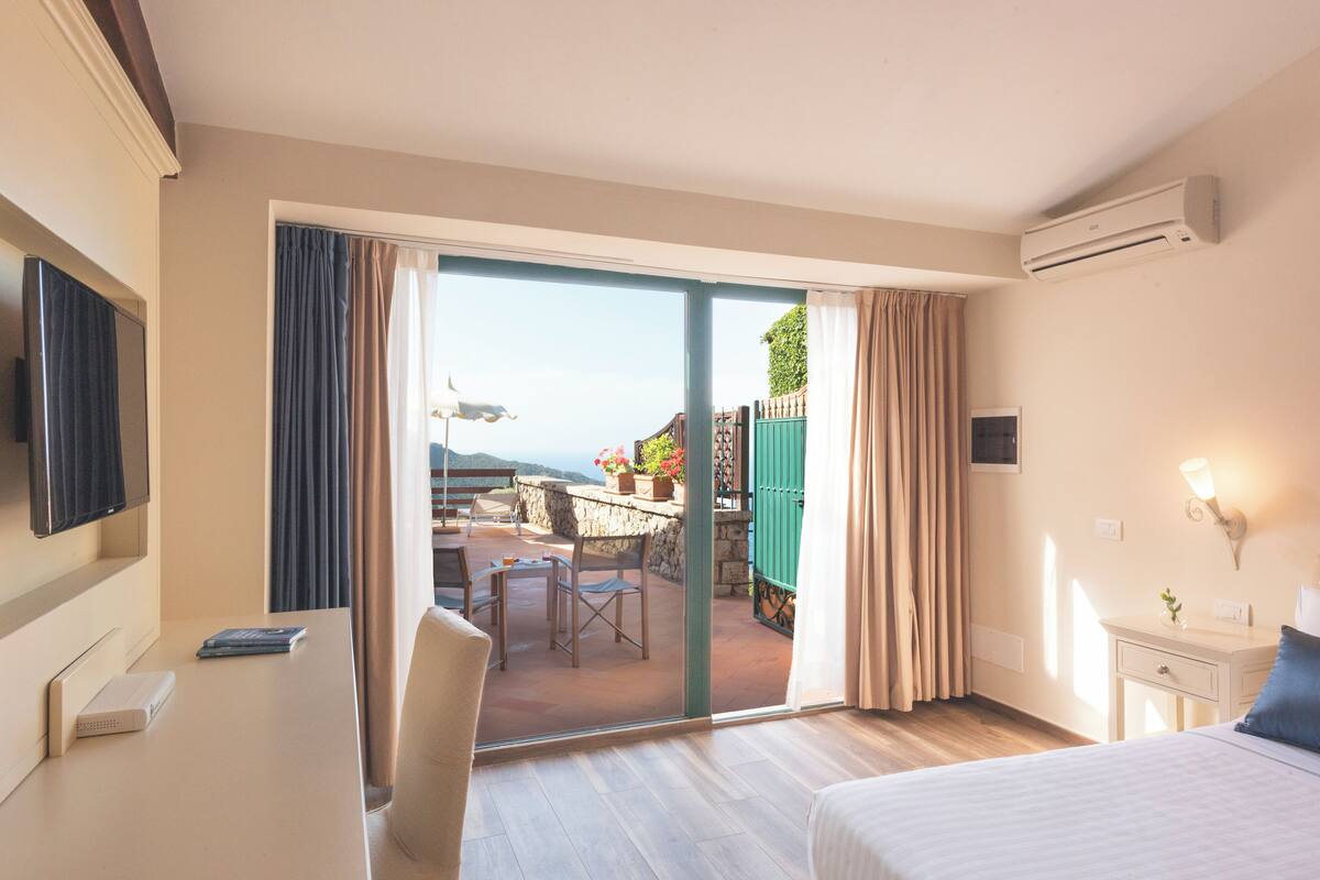 A hotel room with a bed, a desk with a computer, a TV, a sliding glass door to an outdoor area with a table and chairs, and an air conditioning unit.