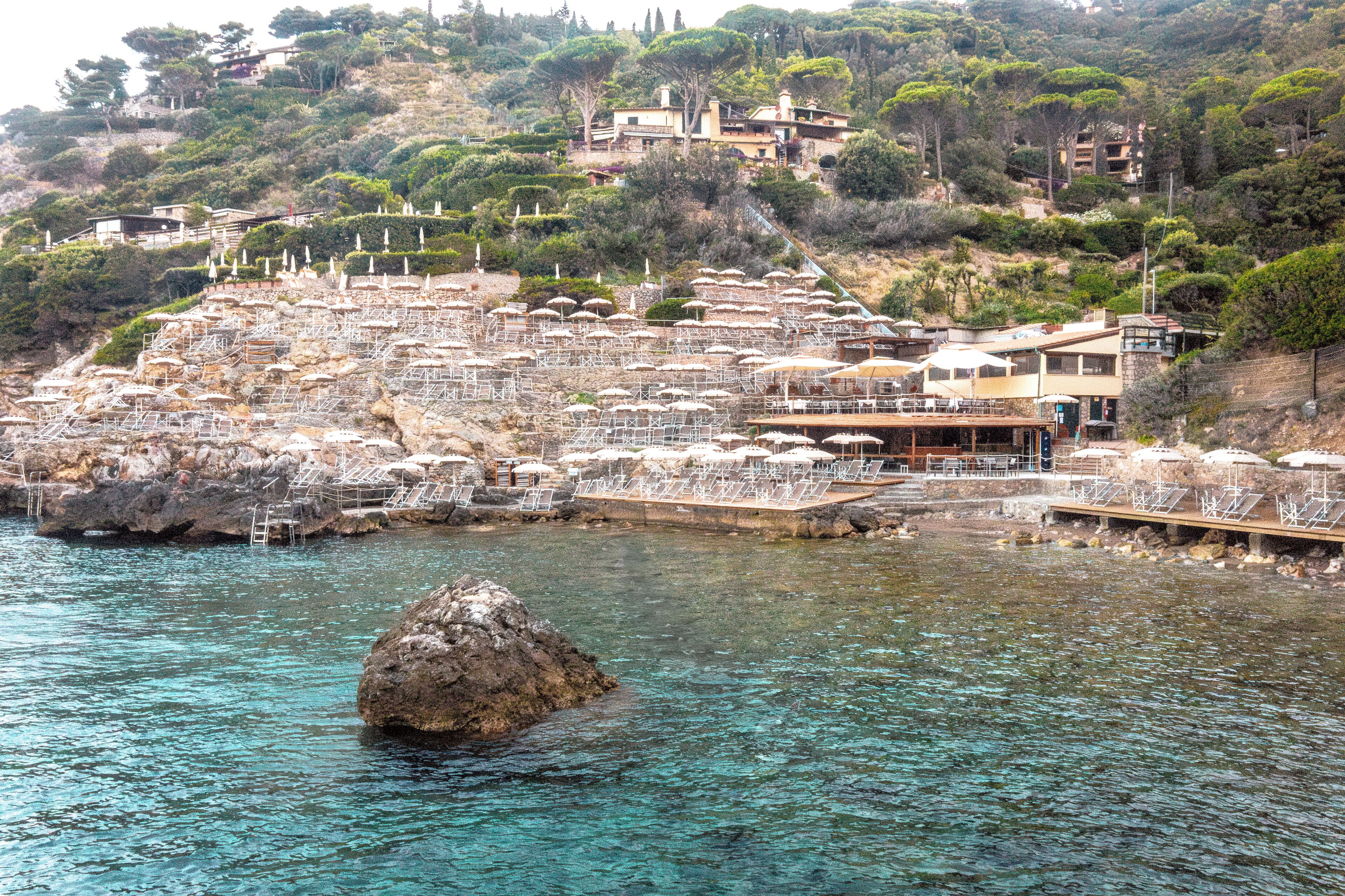 Un hotel costero con varios balcones que dan al mar, una costa rocosa y abundante vegetación en la colina.