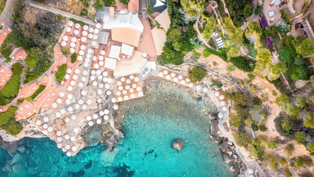 An aerial view of a coastal property with multiple buildings, a beach area, and a rocky shoreline.
