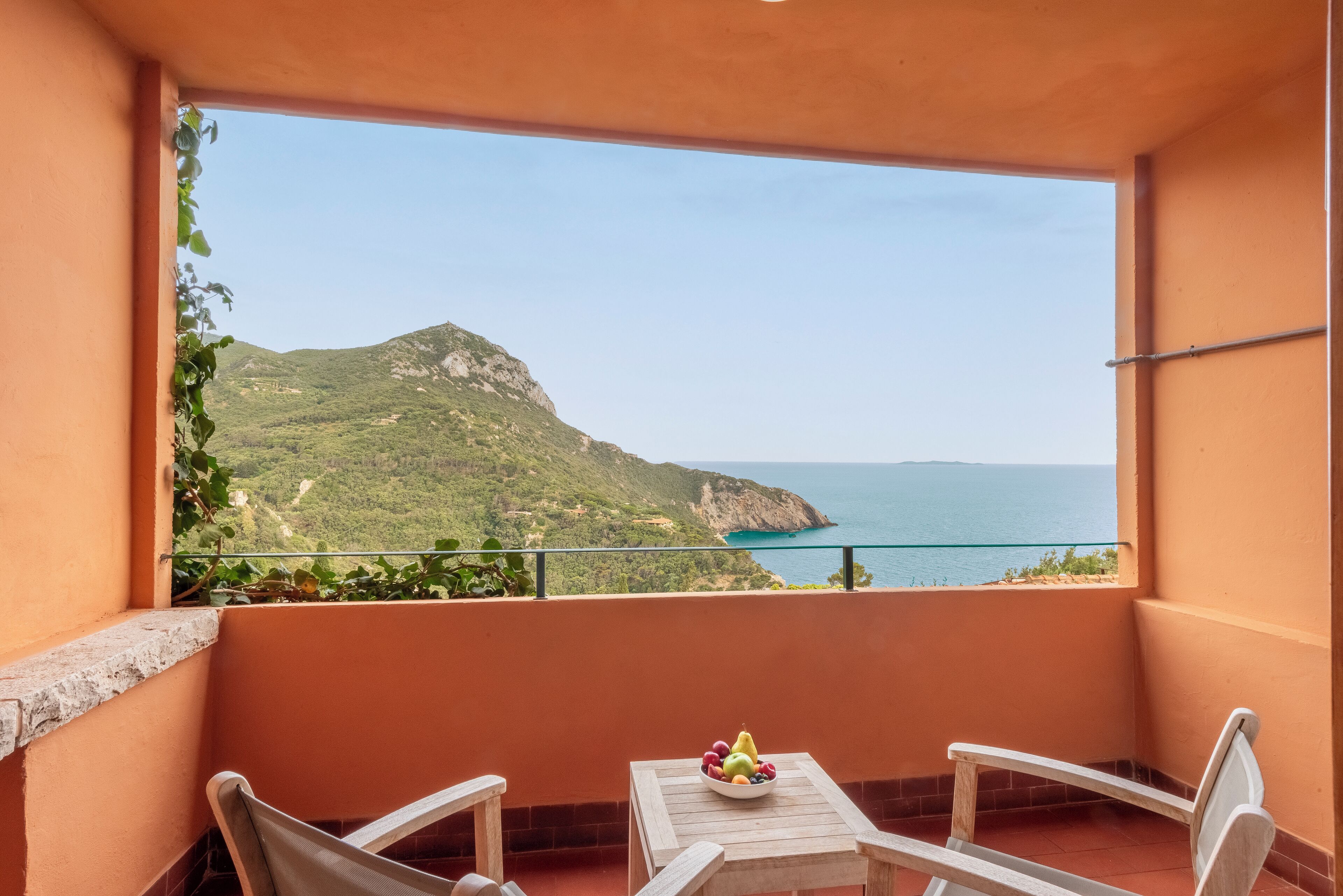 A balcony with a table and chairs, a bowl of fruit, and a view of a coastal landscape.
