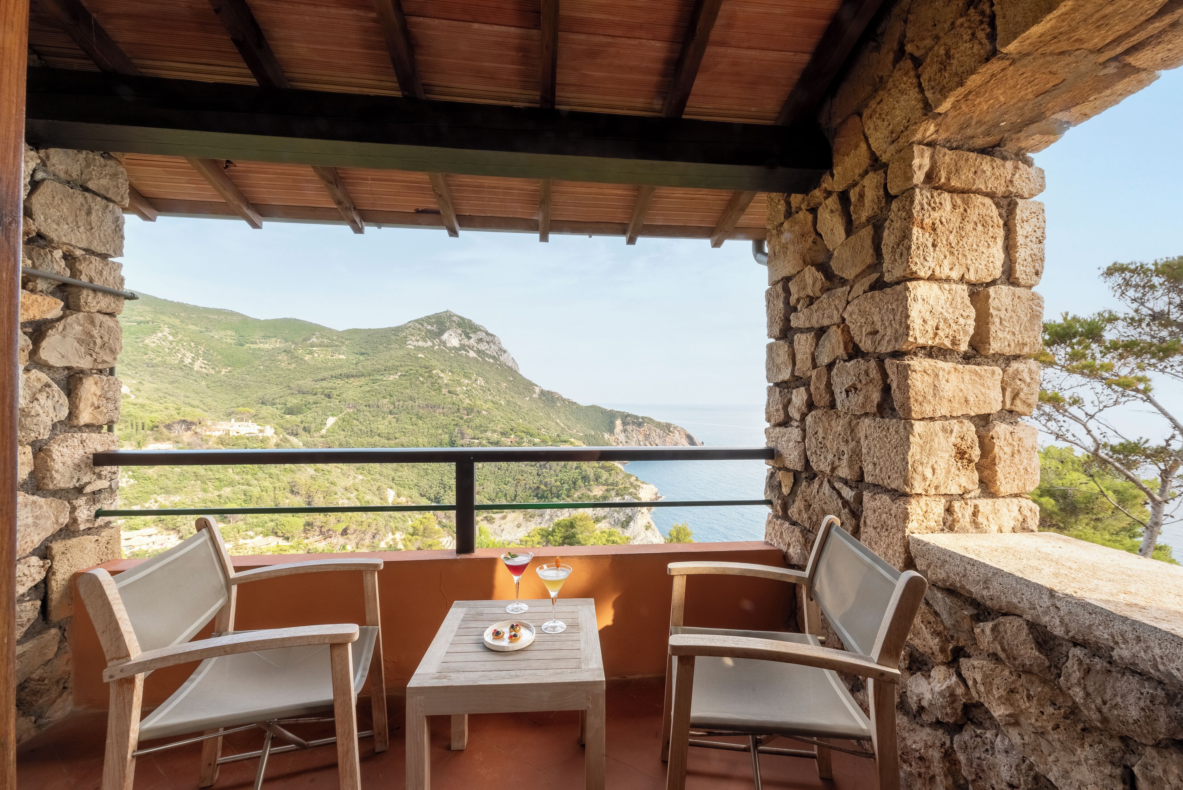 Un balcone con pareti in pietra, due sedie di legno, un tavolino e vista sulle colline verdi e sul mare.