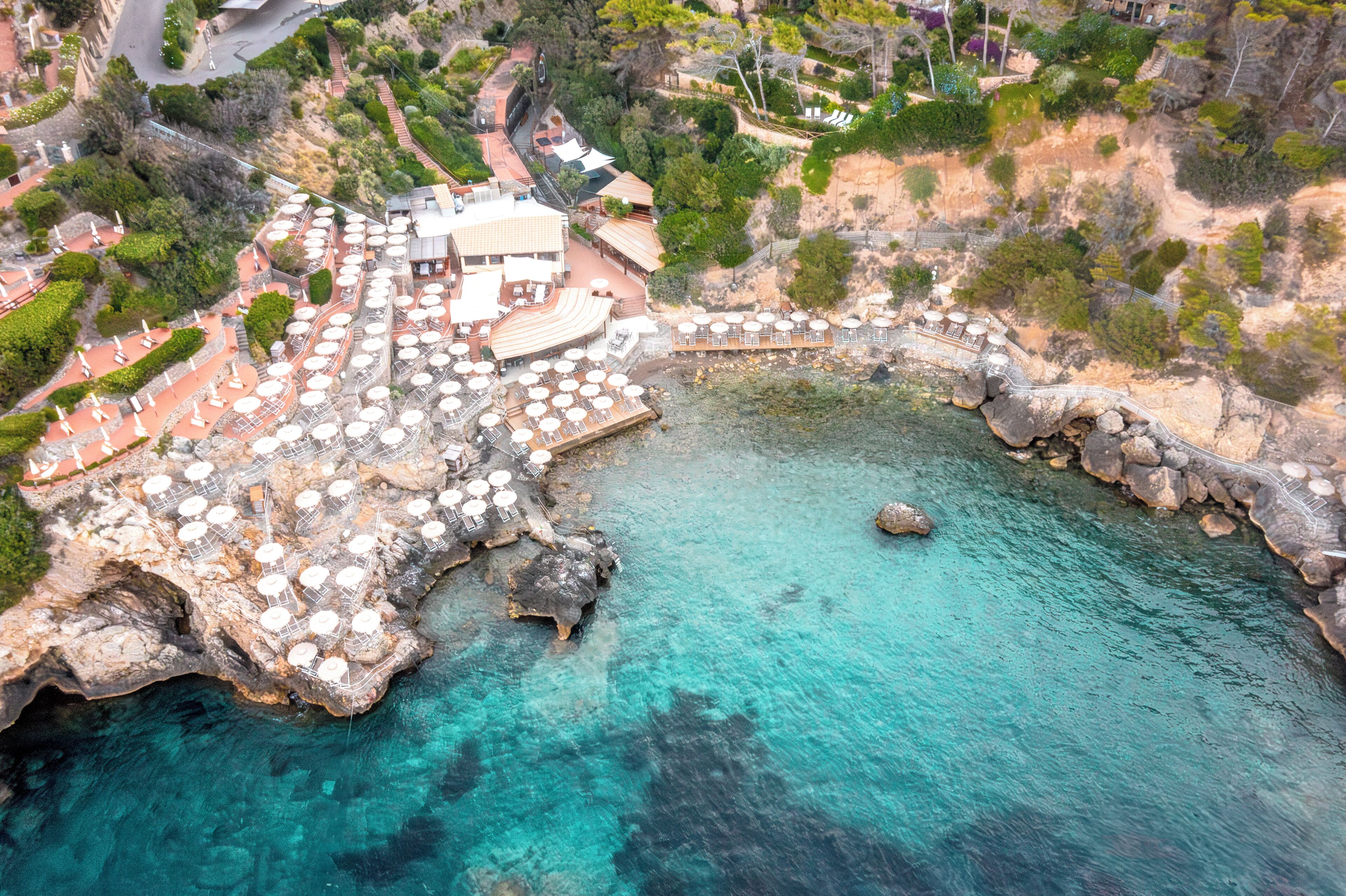 An aerial view of a coastal hotel with outdoor seating areas, a rocky shoreline, and a clear blue sea.