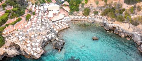 An aerial view of a coastal hotel with outdoor seating areas, a rocky shoreline, and a clear blue sea.