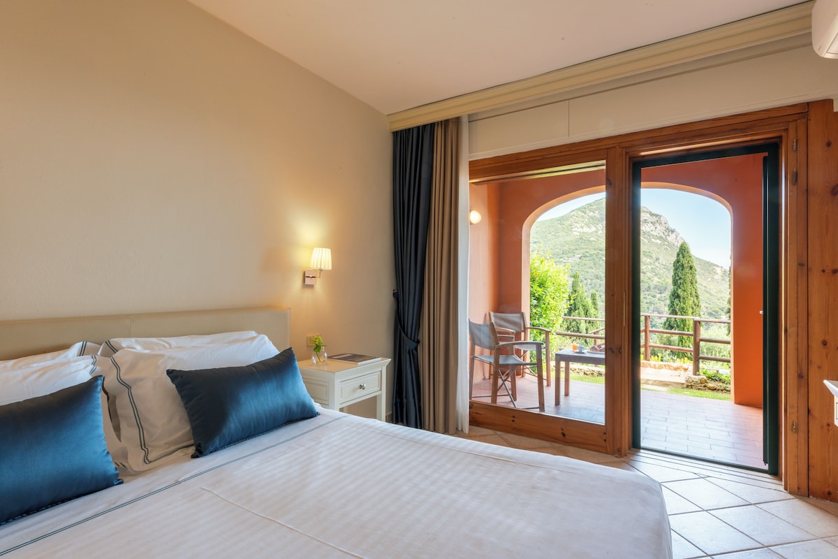 A hotel room with a large bed, a bedside table with a lamp, and a view of a balcony with outdoor furniture.