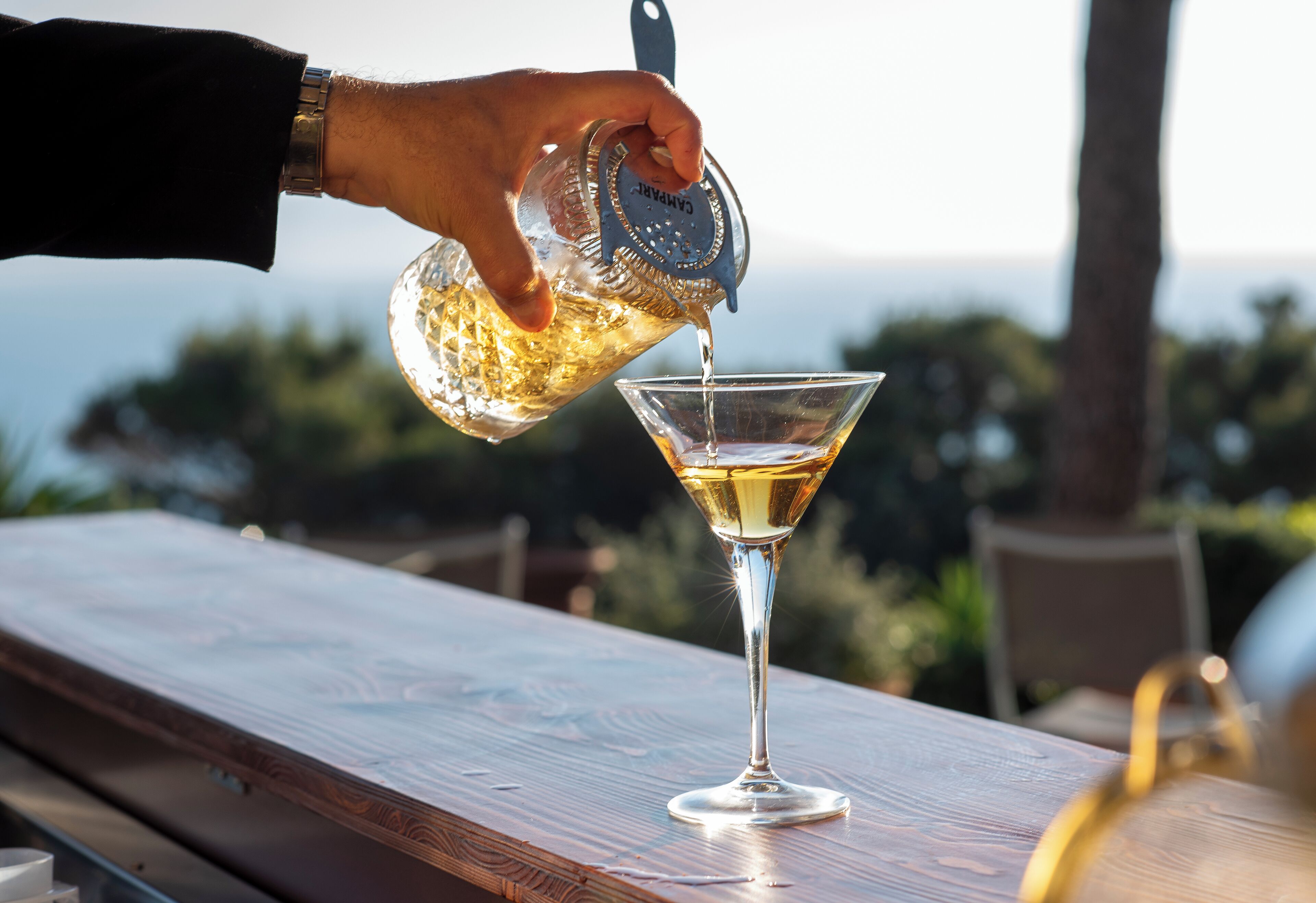 A hand pouring a drink into a martini glass on a wooden table with a view of the ocean in the background.