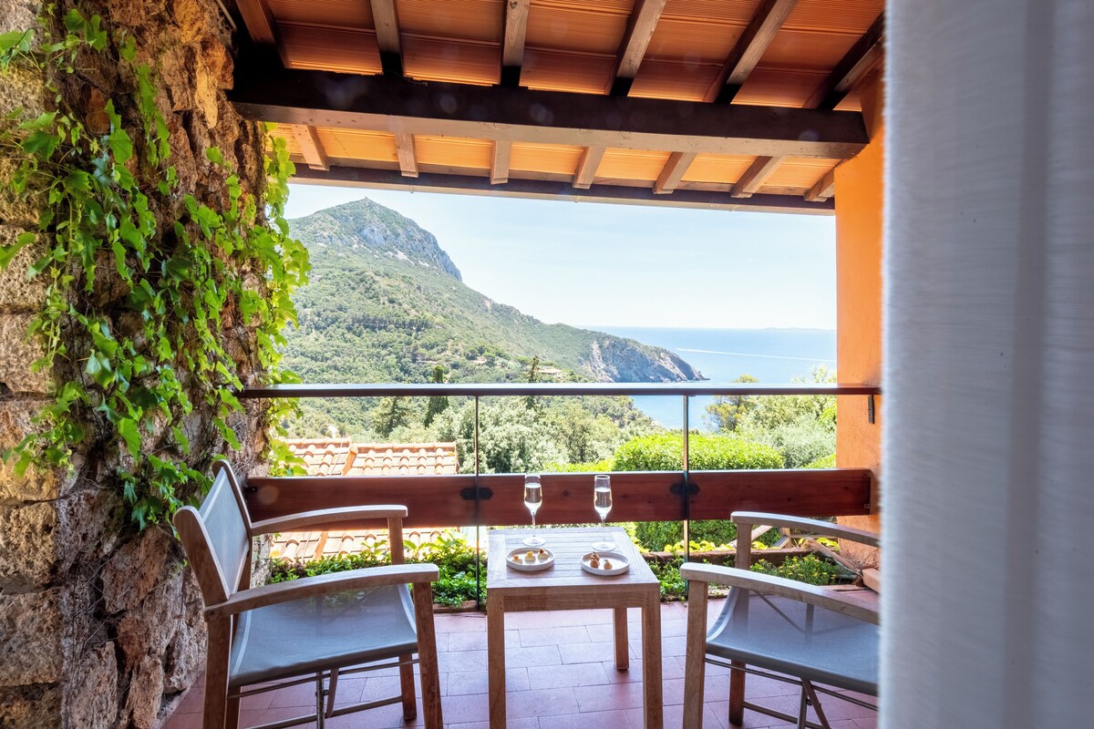 A balcony with a table and chairs, overlooking a mountainous landscape and the sea.