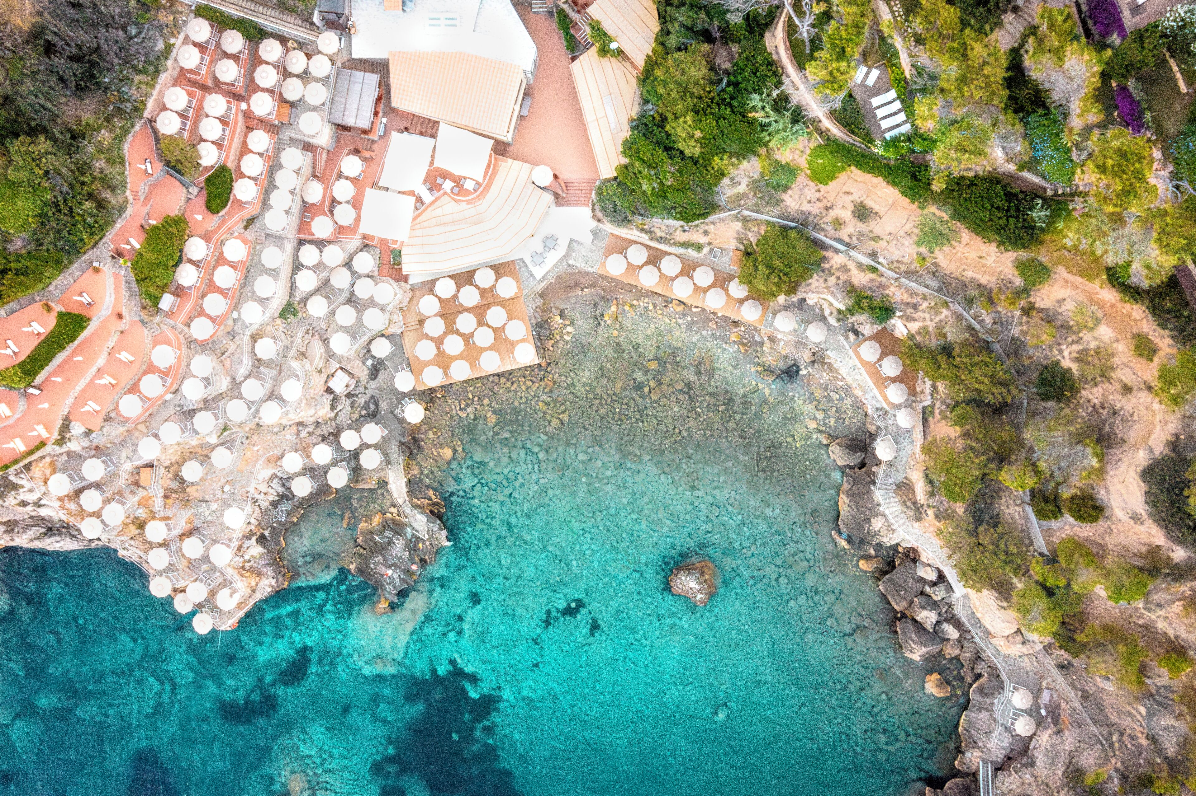 Vista aérea de un complejo turístico costero con varias mesas redondas y sombrillas, un litoral rocoso y un mar azul cristalino.