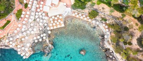 Vista aérea de un complejo turístico costero con varias mesas redondas y sombrillas, un litoral rocoso y un mar azul cristalino.