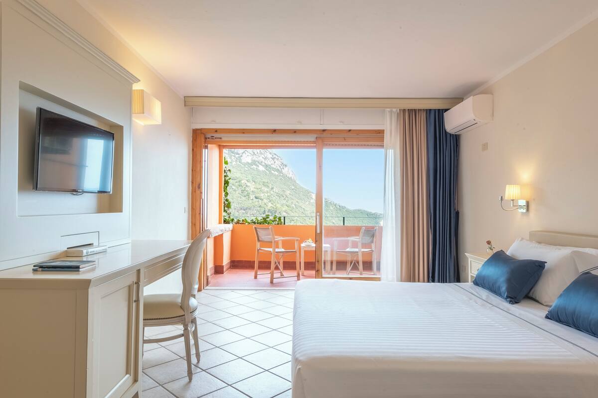 A hotel room with a bed, a desk, a TV, and a balcony with chairs.