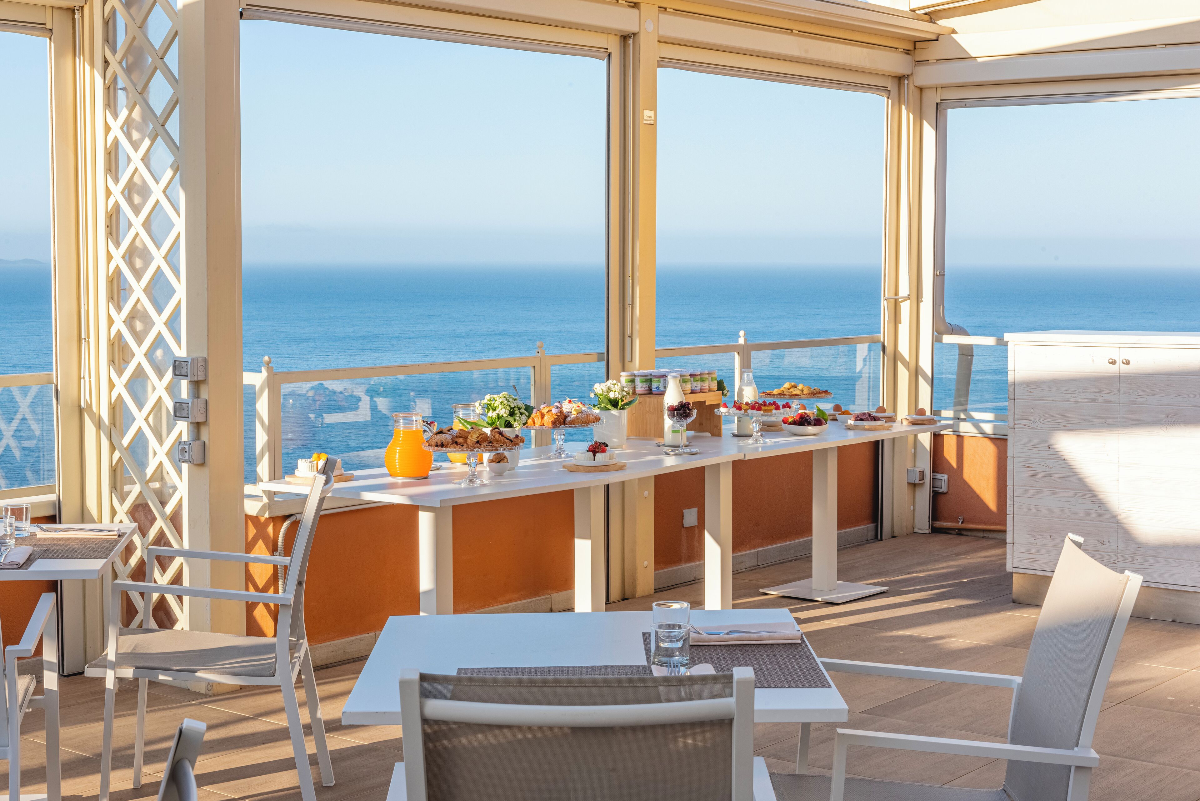 Zona de comedor al aire libre iluminada por el sol, con vistas al mar, mesas con alimentos para el desayuno y una silla blanca.