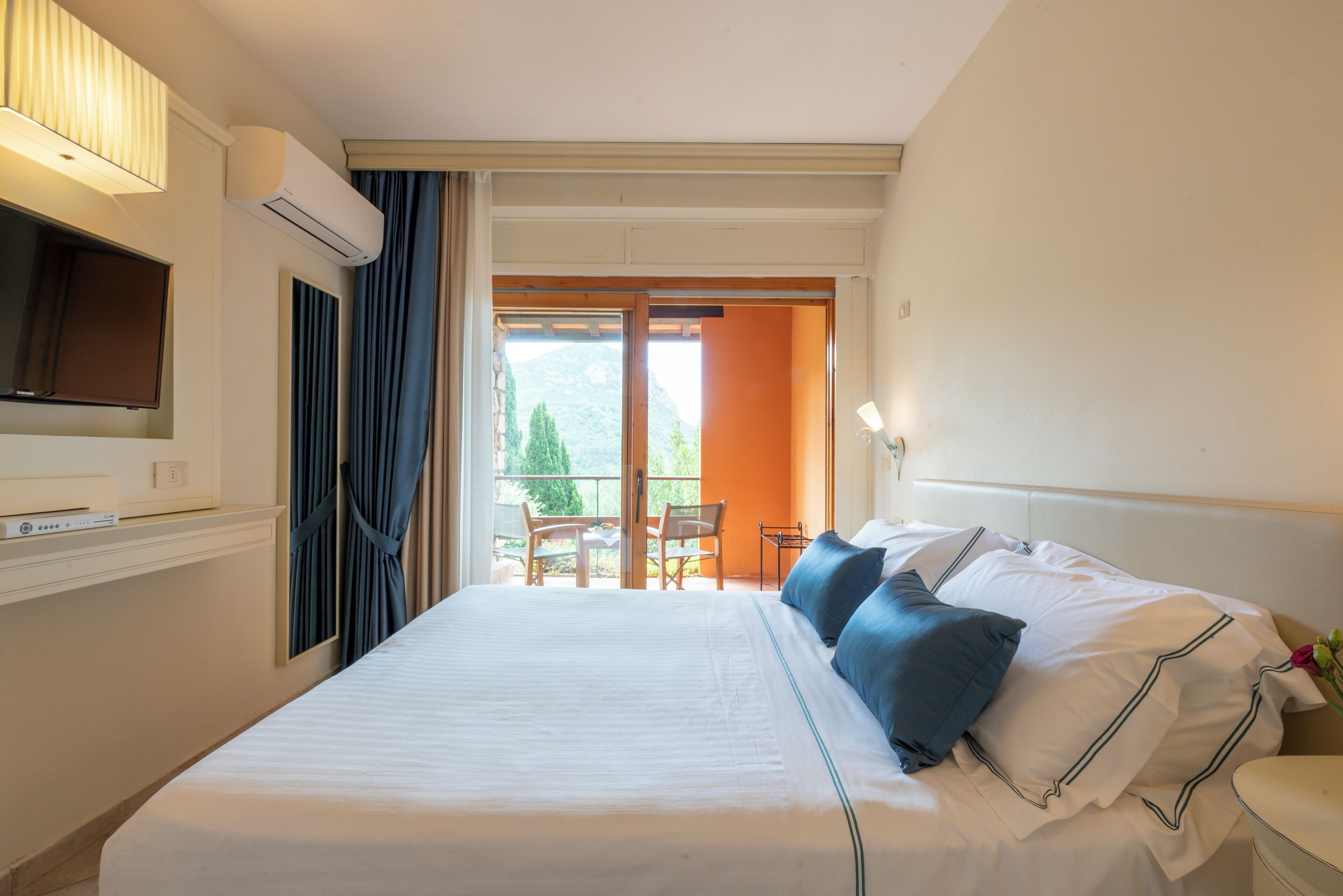 Quarto de hotel com cama, televisão, janela com cortinas e vista para o exterior.