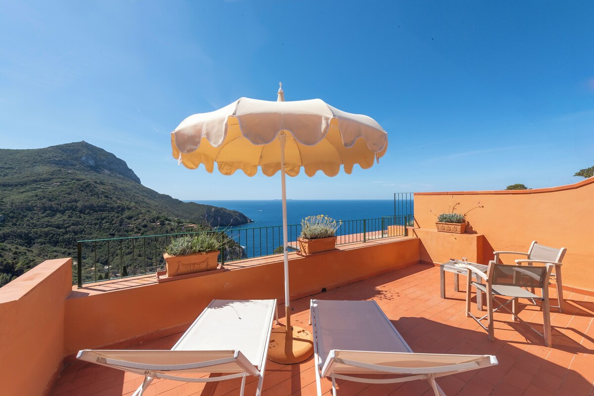 A terrace with a white table and two chairs, a yellow umbrella, and a view of the sea and mountains.