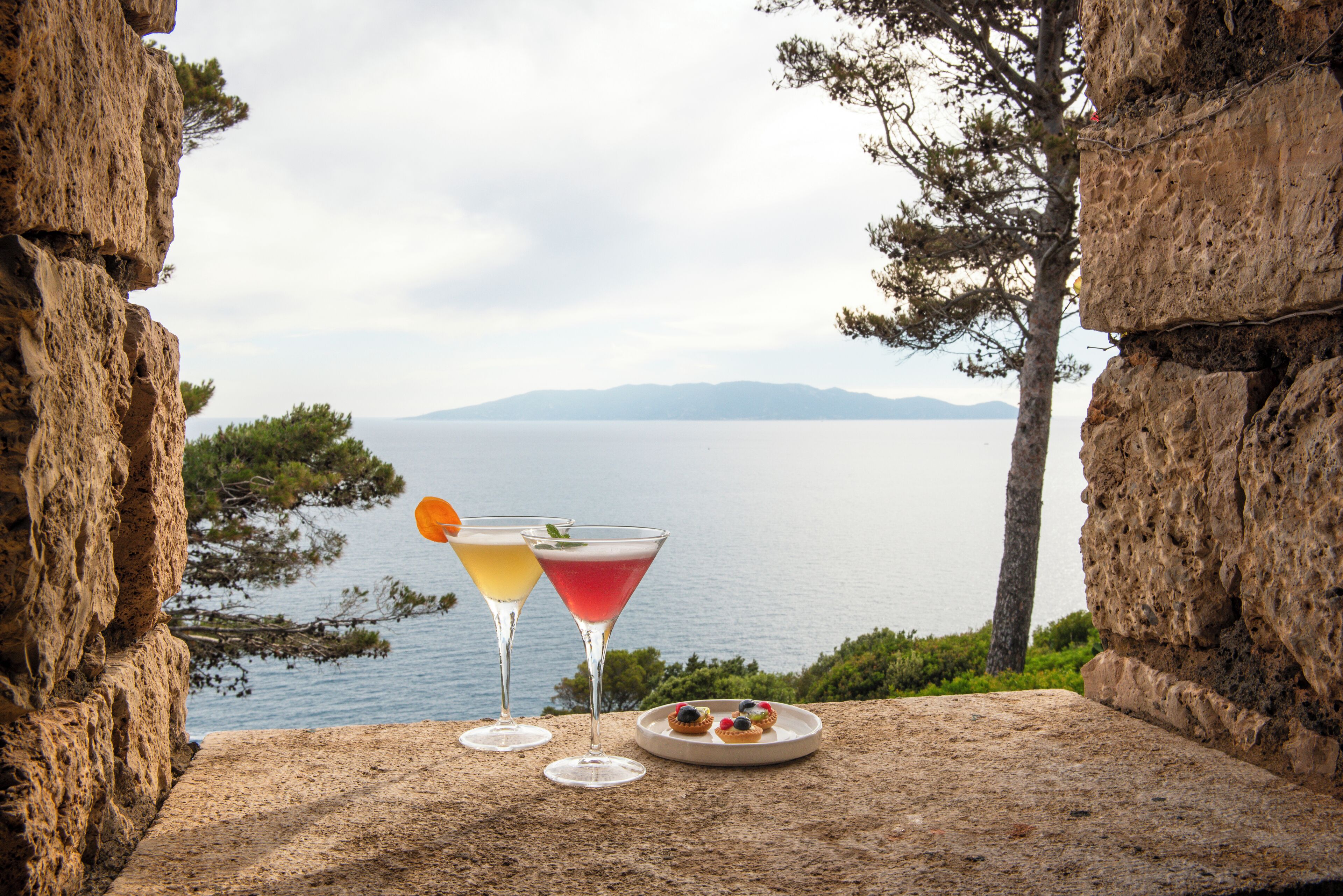 Un tavolo in pietra con due bicchieri di martini e un piatto di pasticcini, con vista su un lago e montagne in lontananza.