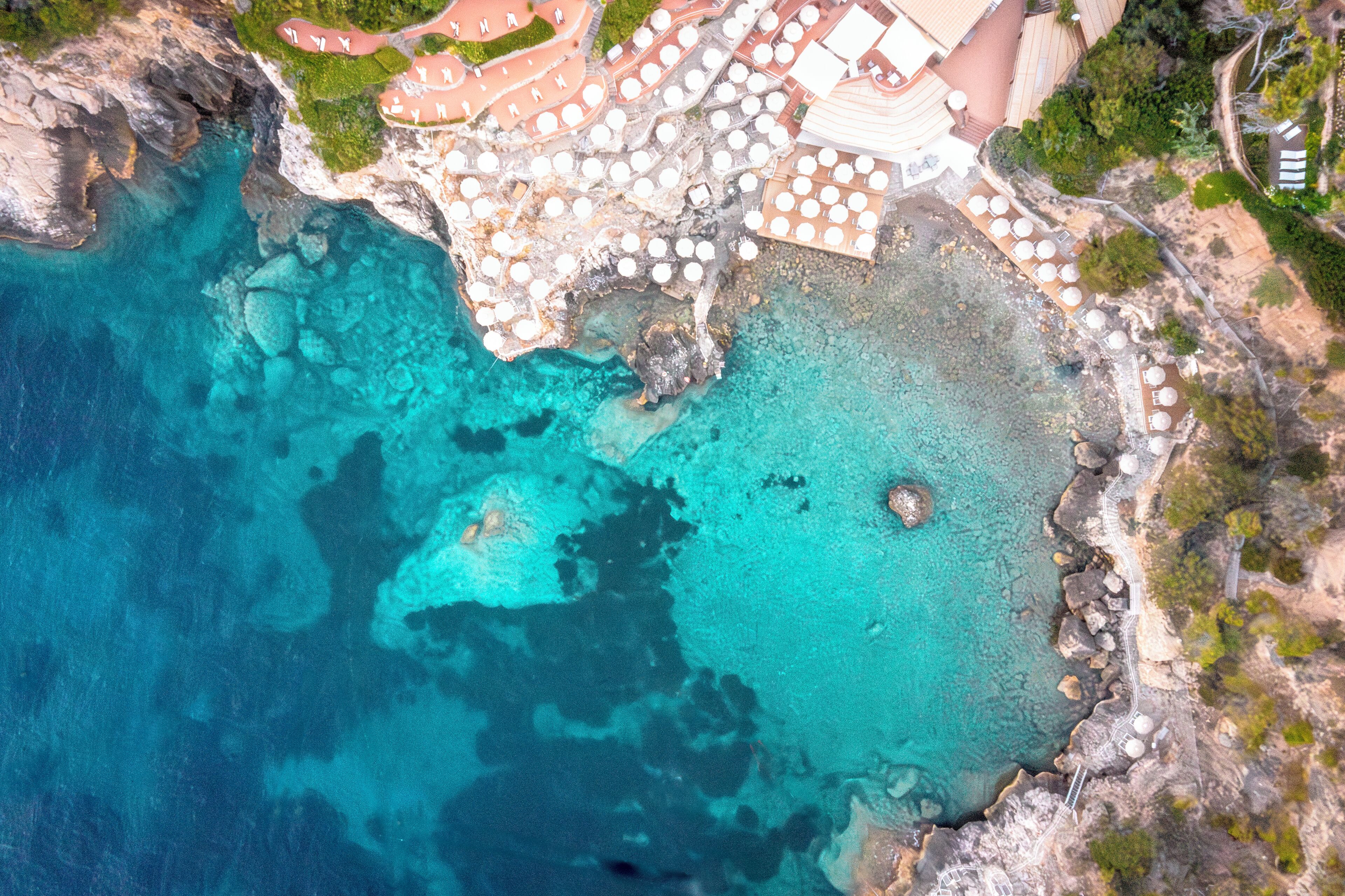 An aerial view of a coastal hotel with a rocky shoreline, clear turquoise waters, and numerous umbrellas.