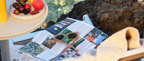 Une table sur laquelle se trouvent un plateau de fruits, une boisson et des lunettes de soleil posées sur un magazine, le tout installé sur une chaise avec une serviette.