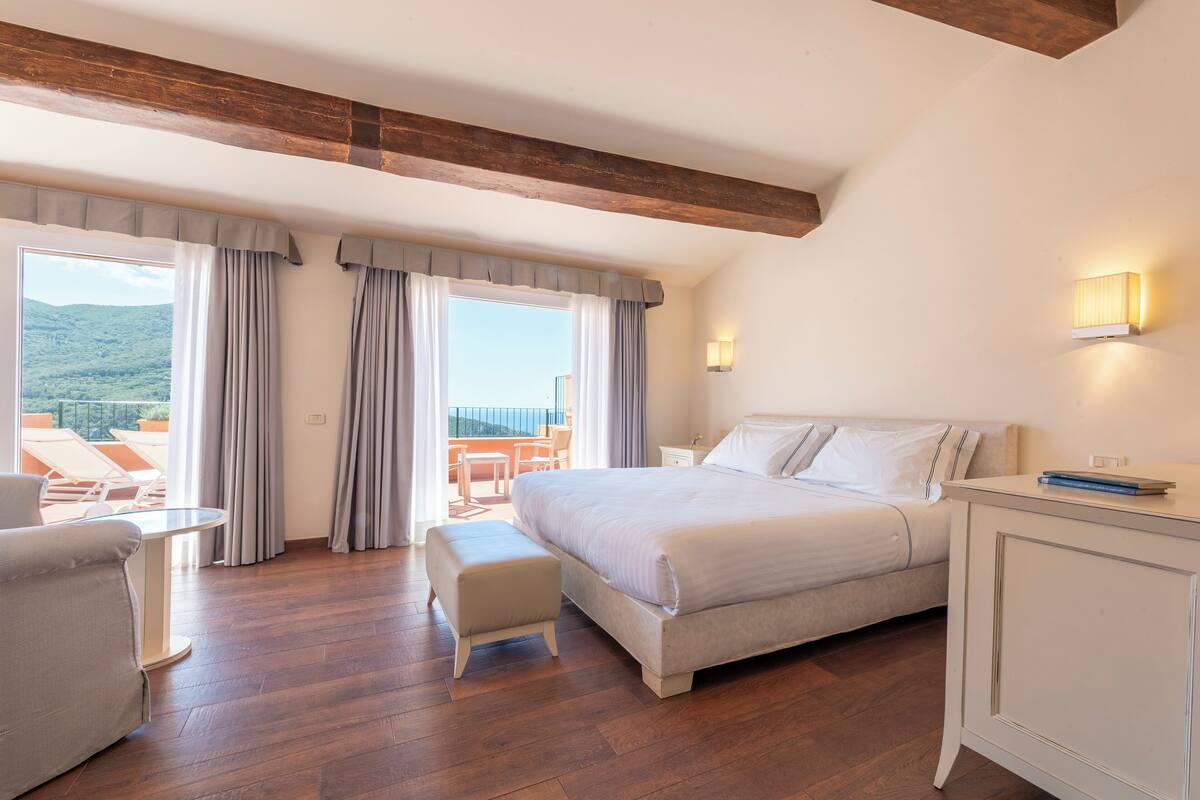 A bedroom with a large bed, a small table, a chair, and a view of the outdoors.