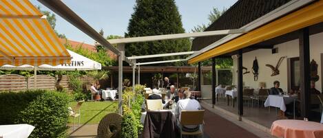 An outdoor patio area with tables and chairs, a yellow awning, and a sign that reads "Veltins."