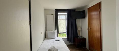 Ein Hotelzimmer mit einem Einzelbett, einem an der Wand montierten Flachbildfernseher, einer Holztür und einem Fenster mit Vorhängen.