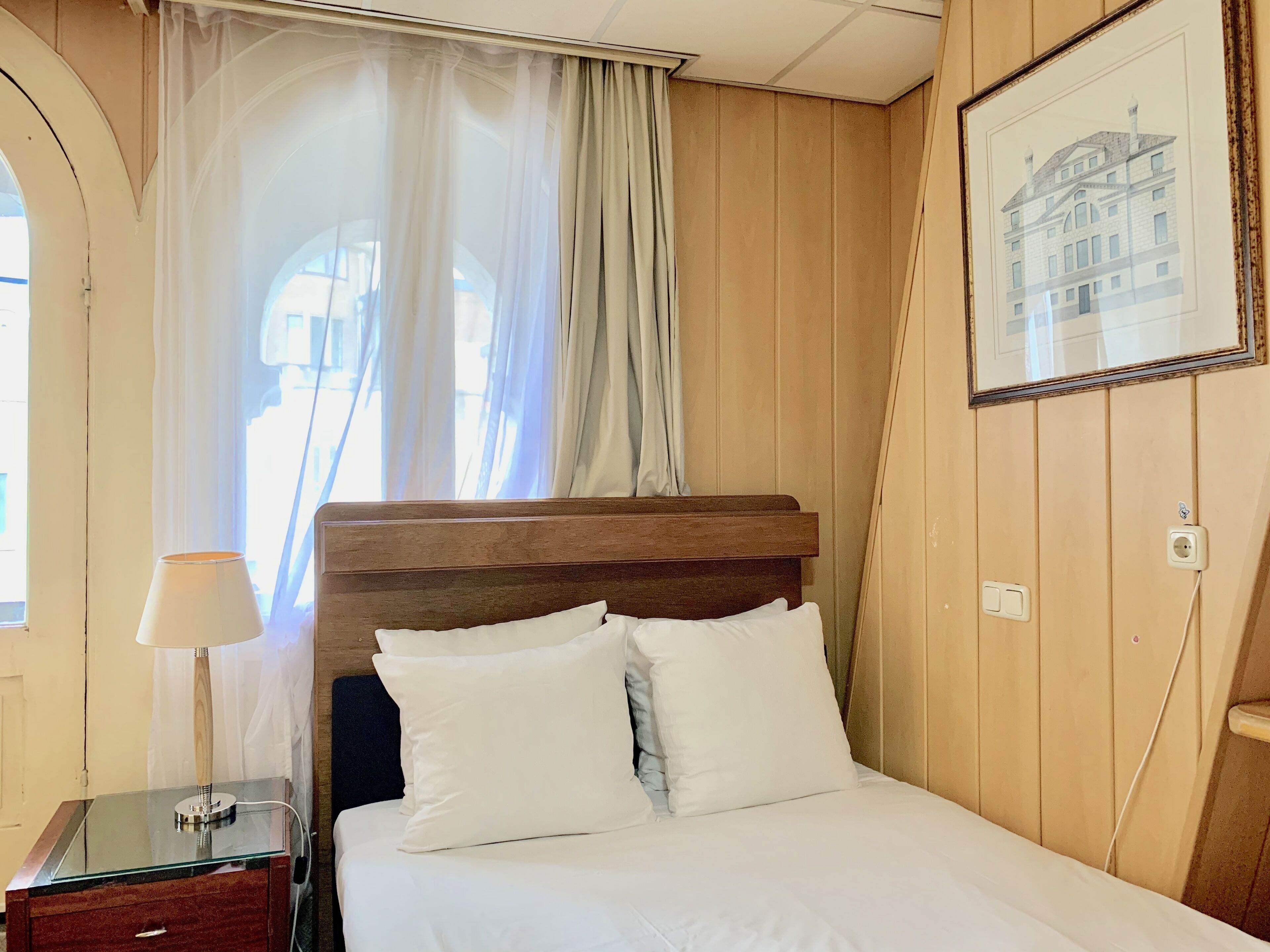 Un lit bien fait, avec du linge de lit blanc et une tête de lit en bois, une lampe de chevet et un tableau encadré au mur.