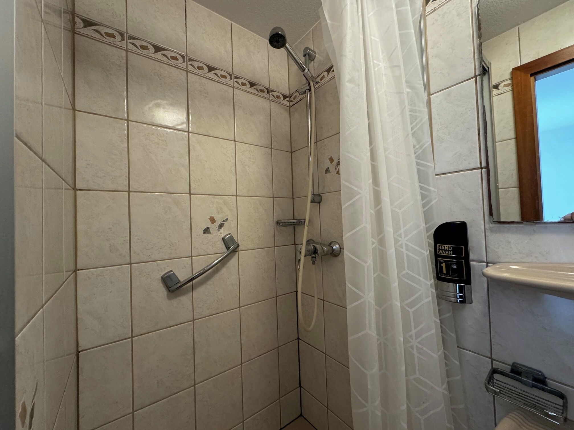 Ein Badezimmer mit weiß gefliesten Wänden, einer Dusche inklusive Handbrause, einem Waschbecken und einem Spiegel mit Holzrahmen.