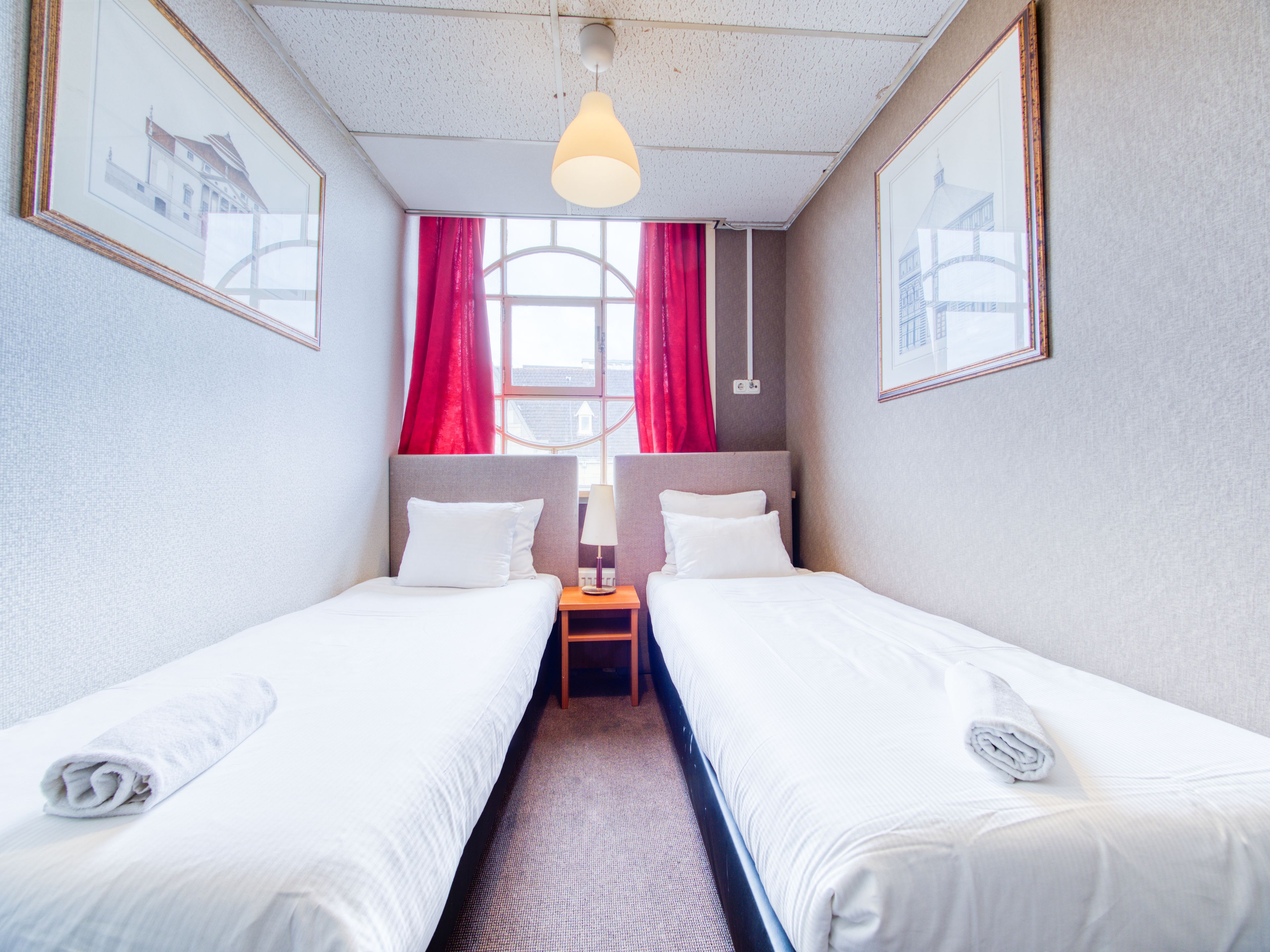 A hotel room with two single beds, a small table, a lamp, and a window with red curtains.