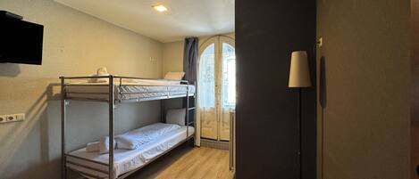 A room with a bunk bed, a flat bed, a TV mounted on the wall, a lamp, and a window with curtains.